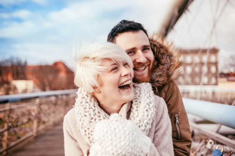 Lachendes Pärchen in Winterkleidung steht auf einer Brücke und umarmt sich