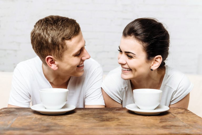 Mann und Frau sitzen mit Kaffeetassen vor sich nebeneinander am Tisch und lächeln sich an