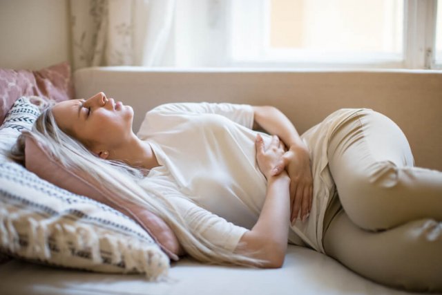 Eine Frau liegt mit schmerzverzerrtem Gesicht auf dem Sofa vor einem Fenster und presst die Hände auf ihren Bauch