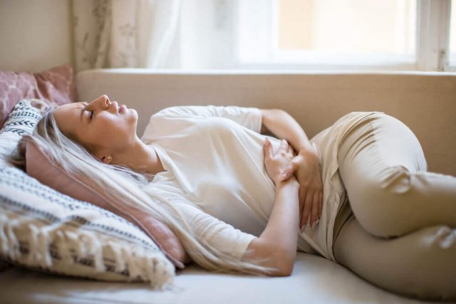 Eine Frau liegt mit schmerzverzerrtem Gesicht auf dem Sofa vor einem Fenster und presst die Hände auf ihren Bauch