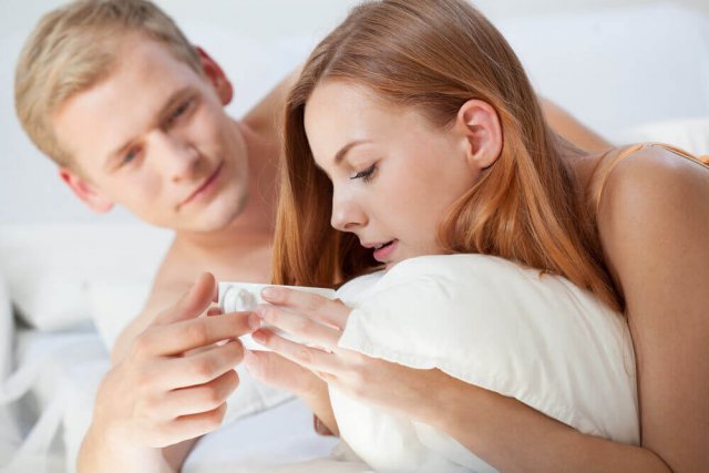 Ein Paar liegt im Bett, sie liegt auf dem Bauch und hält eine Kaffeetasse, er berührt ihre Hand