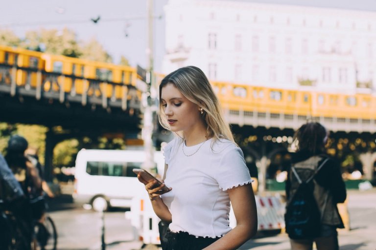 Eine blonde Frau mit weißem T-Shirt schaut auf ihr Handy, während sie die Straße hinuntergeht