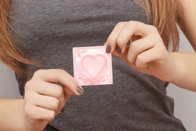 Eine junge Frau mit dunkel lackierten Nägeln hält eine Kondomverpackung mit Herz zwischen den Fingern