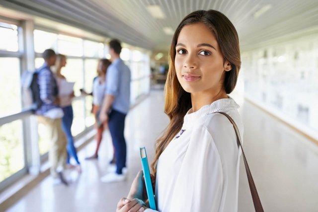 Eine junge Frau mit türkisem Notizbuch schaut in die Kamera, eine Gruppe steht dahinter im Gang am Fenster